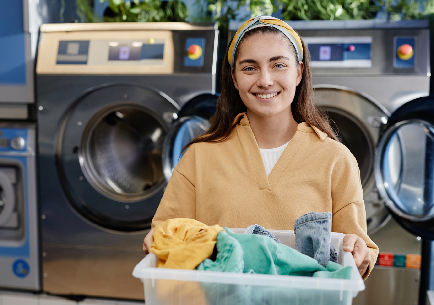 A woman smiles while holding a laundry basket with clothes. Behind her are washing machines in a laundromat.