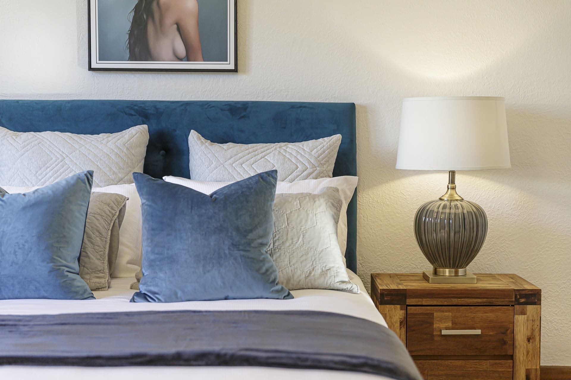 Bedroom with a blue headboard bed, pillows, a bedside table, and lamp. Wall artwork is partially visible above the bed.
