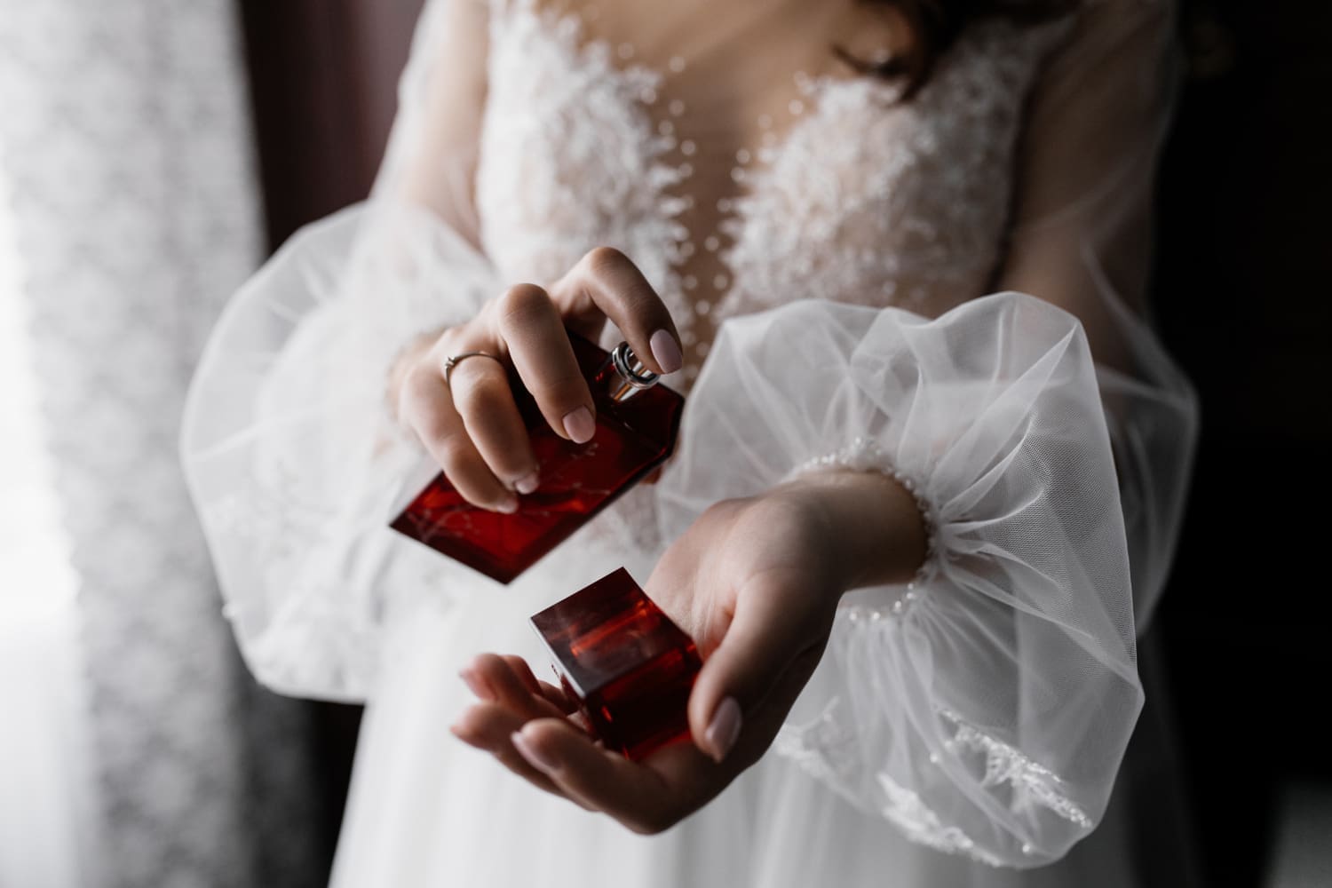 Person in a lace dress applying perfume from a red bottle onto their wrist.