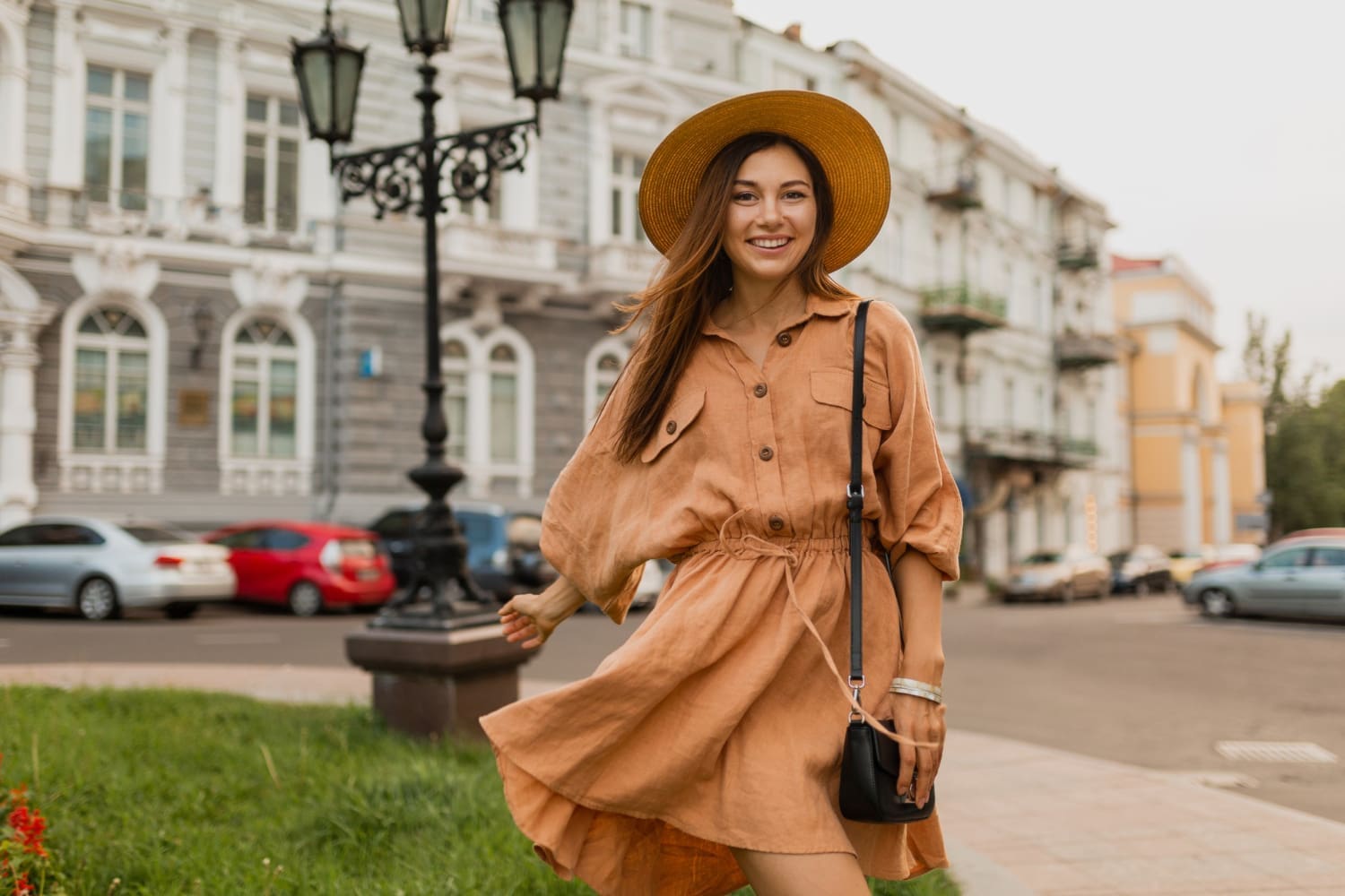 Woman in a brown dress and straw hat smiles while standing on a street with historic buildings and cars in the background.