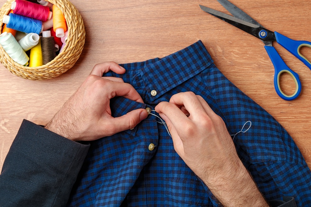 A person sewing a button onto a blue plaid shirt, with scissors and a basket of colorful spools of thread on a wooden table.