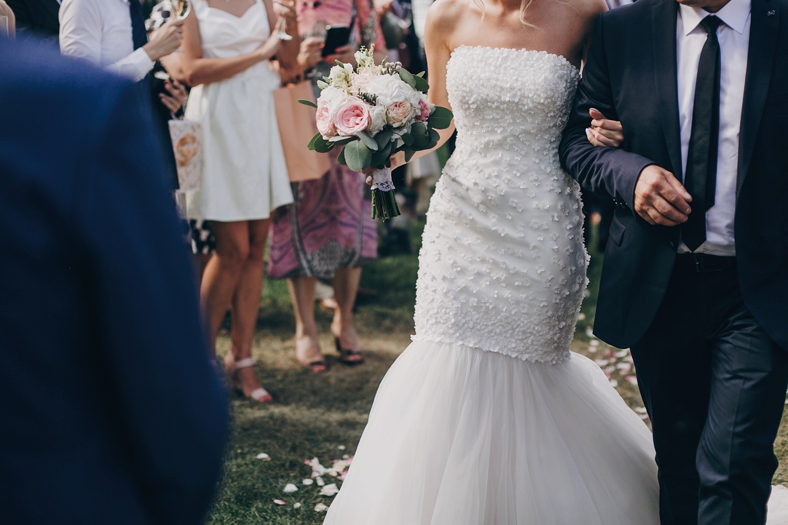 A bride in a white strapless wedding dress holding a bouquet walks arm in arm with a man in a suit at an outdoor ceremony, surrounded by guests.