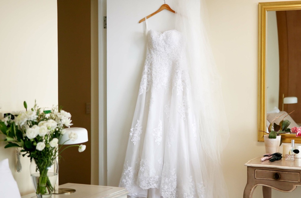 A white lace wedding dress hangs on a door next to a mirror, with a bouquet of white flowers and makeup items on a nearby vanity table.