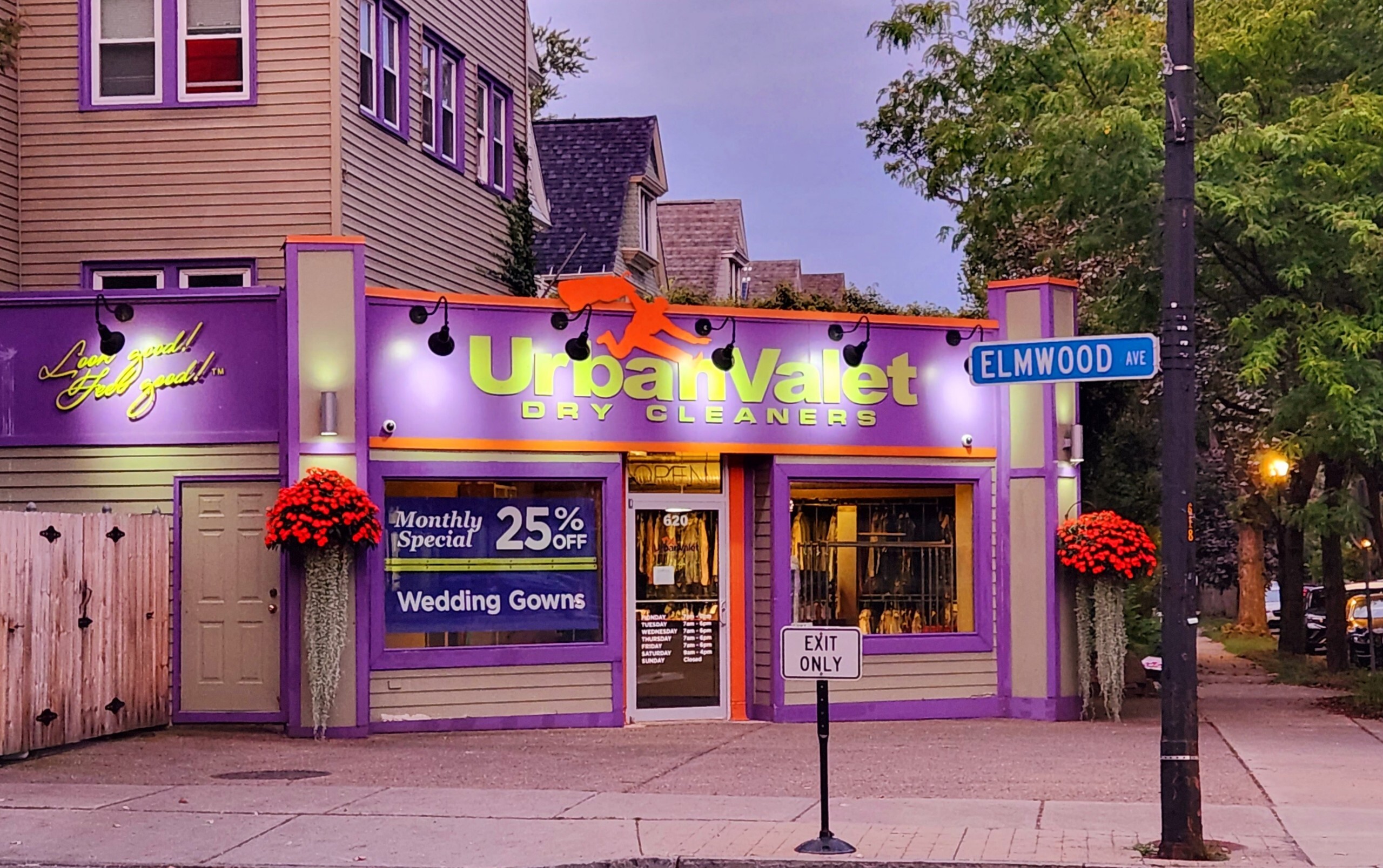 A purple storefront for Urban Valet Dry Cleaners is at the corner of Elmwood Ave, with signs advertising a 25% off special and wedding gown cleaning. Red flower baskets decorate the front.