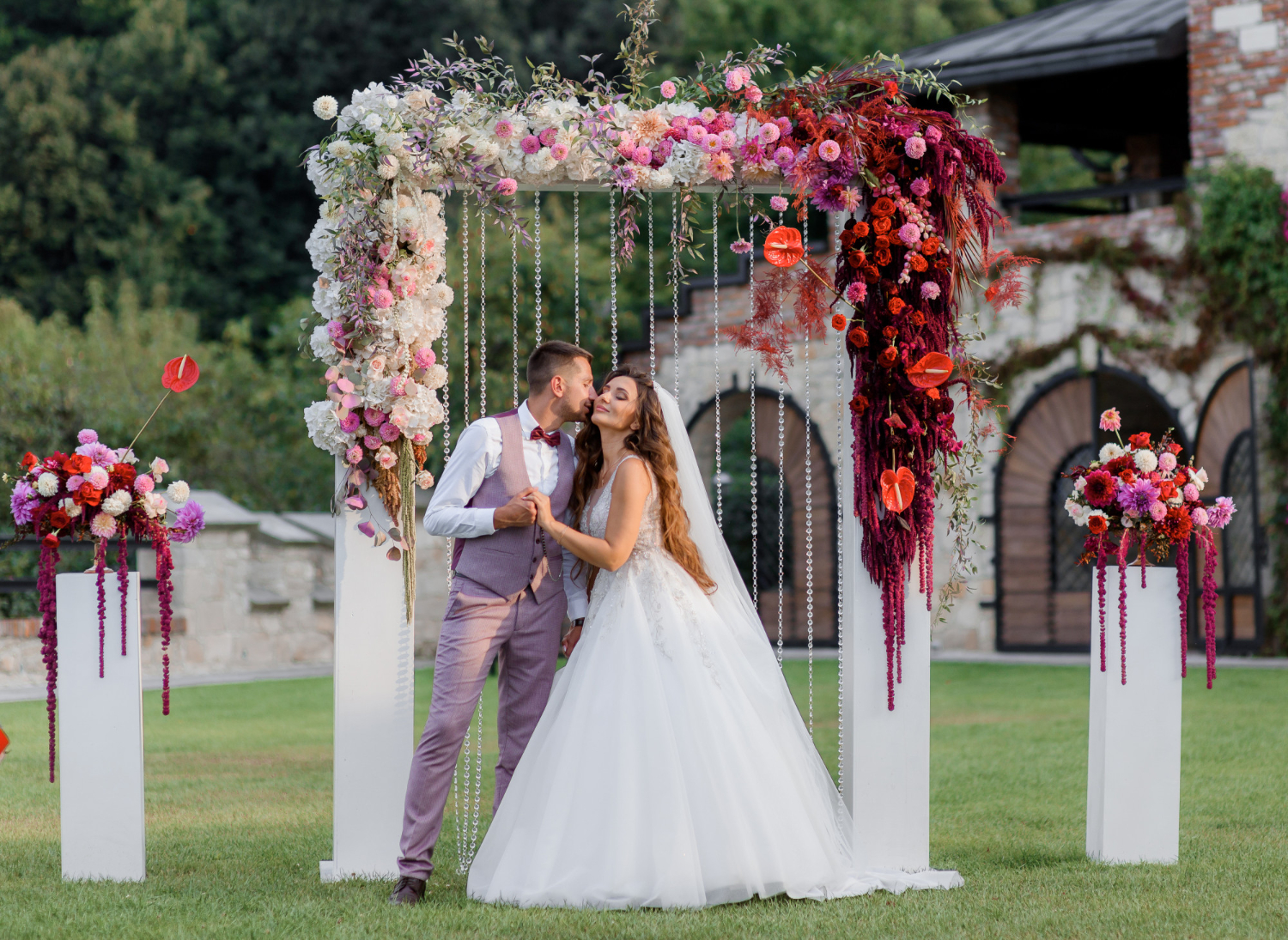A bride and groom kiss under a floral wedding arch outdoors, surrounded by colorful flower arrangements on a grassy lawn.