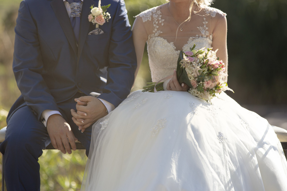 A bride in a white lace wedding dress and a groom in a dark blue suit sit side by side; the bride holds a bouquet of flowers.