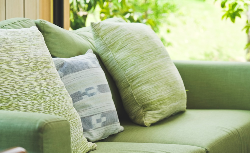 A green sofa with three cushions, two in a matching green fabric and one in a light grey pattern, positioned near a window with greenery outside.