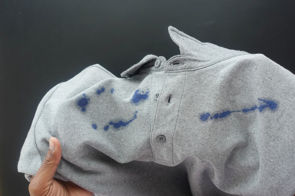 A hand holds a grey shirt with blue ink stains on the sleeve and chest area against a dark background.