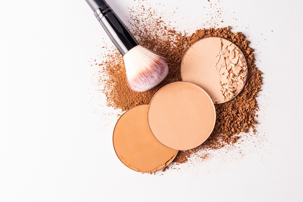 A makeup brush rests on three opened powder compacts in various shades, with powder scattered on a white surface.