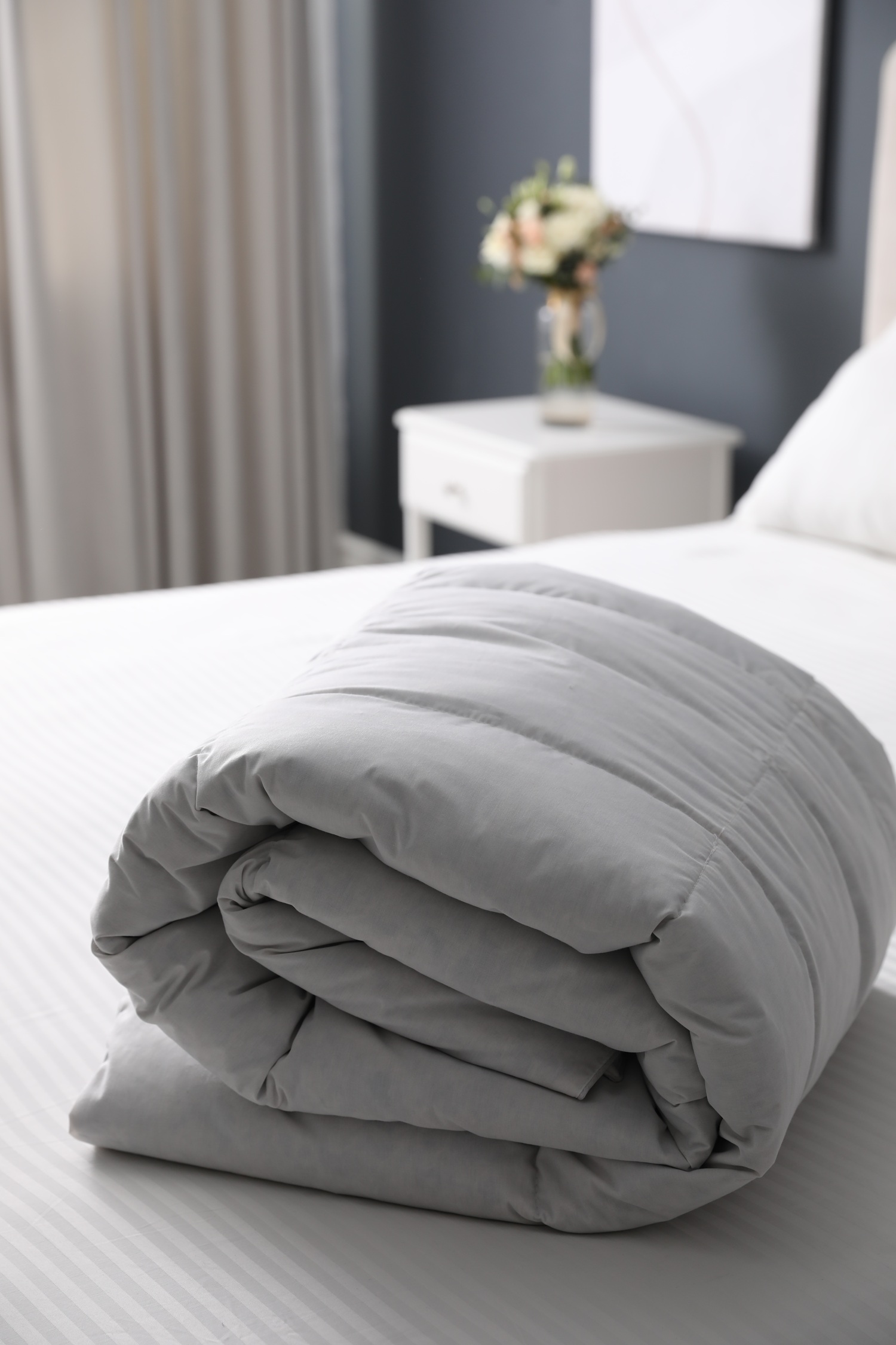 A neatly rolled light gray comforter sits on a white-striped bed in a modern bedroom with a nightstand and a vase of flowers in the background. A neatly rolled light gray comforter sits on a white-striped bed in a modern bedroom with a nightstand and a vase of flowers in the background.