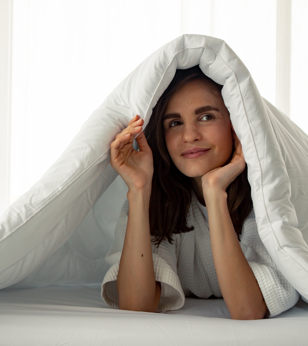 A woman in a white robe smiles while lying under a white comforter, with the blanket draped over her head. A woman in a white robe smiles while lying under a white comforter, with the blanket draped over her head.