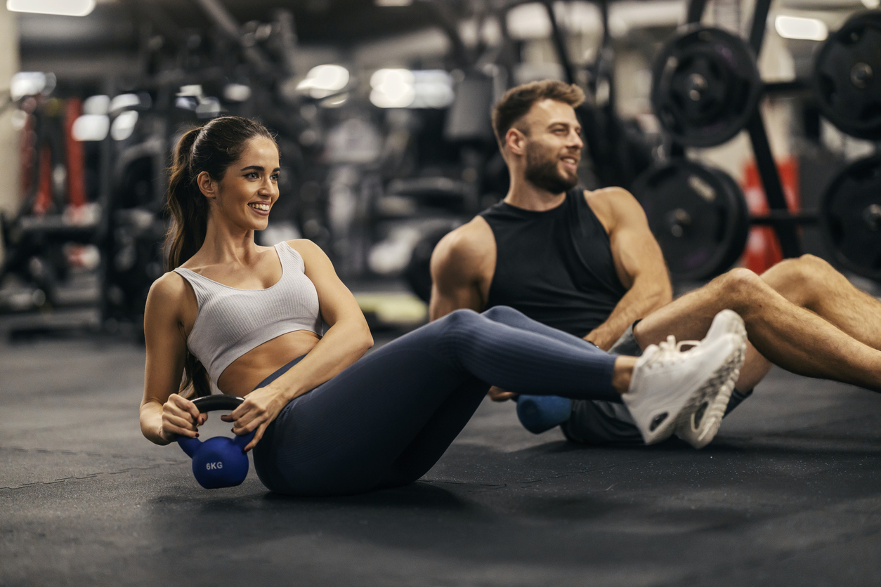 Two people exercise on a gym floor, performing Russian twists with kettlebells. Gym equipment is visible in the background.