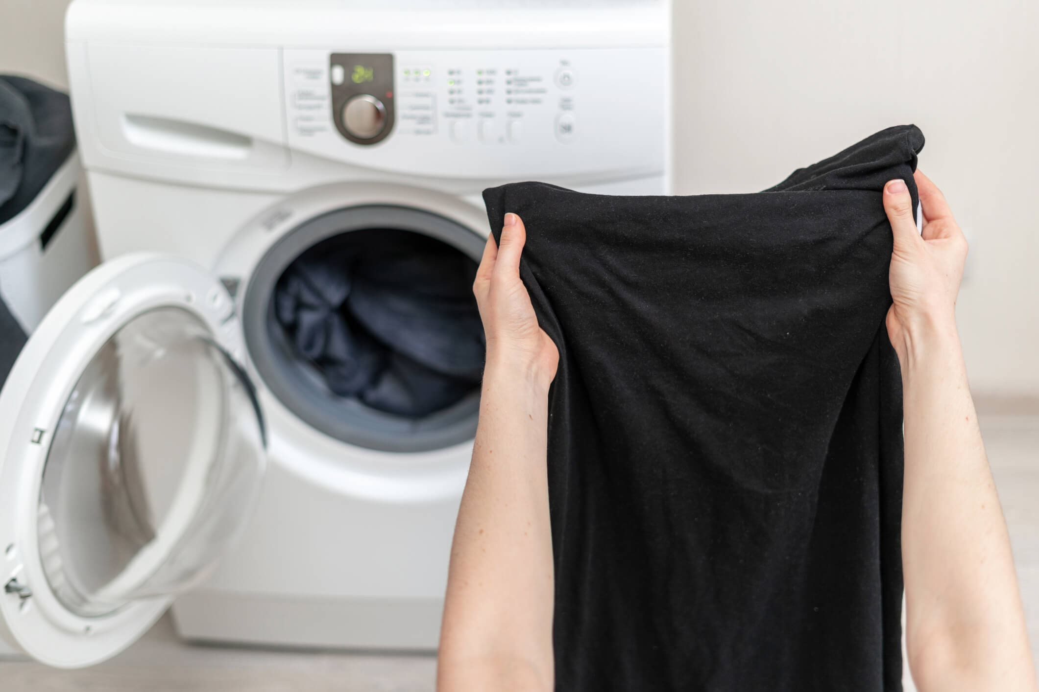 A person holds up a black garment in front of an open front-loading washing machine. A person holds up a black garment in front of an open front-loading washing machine.